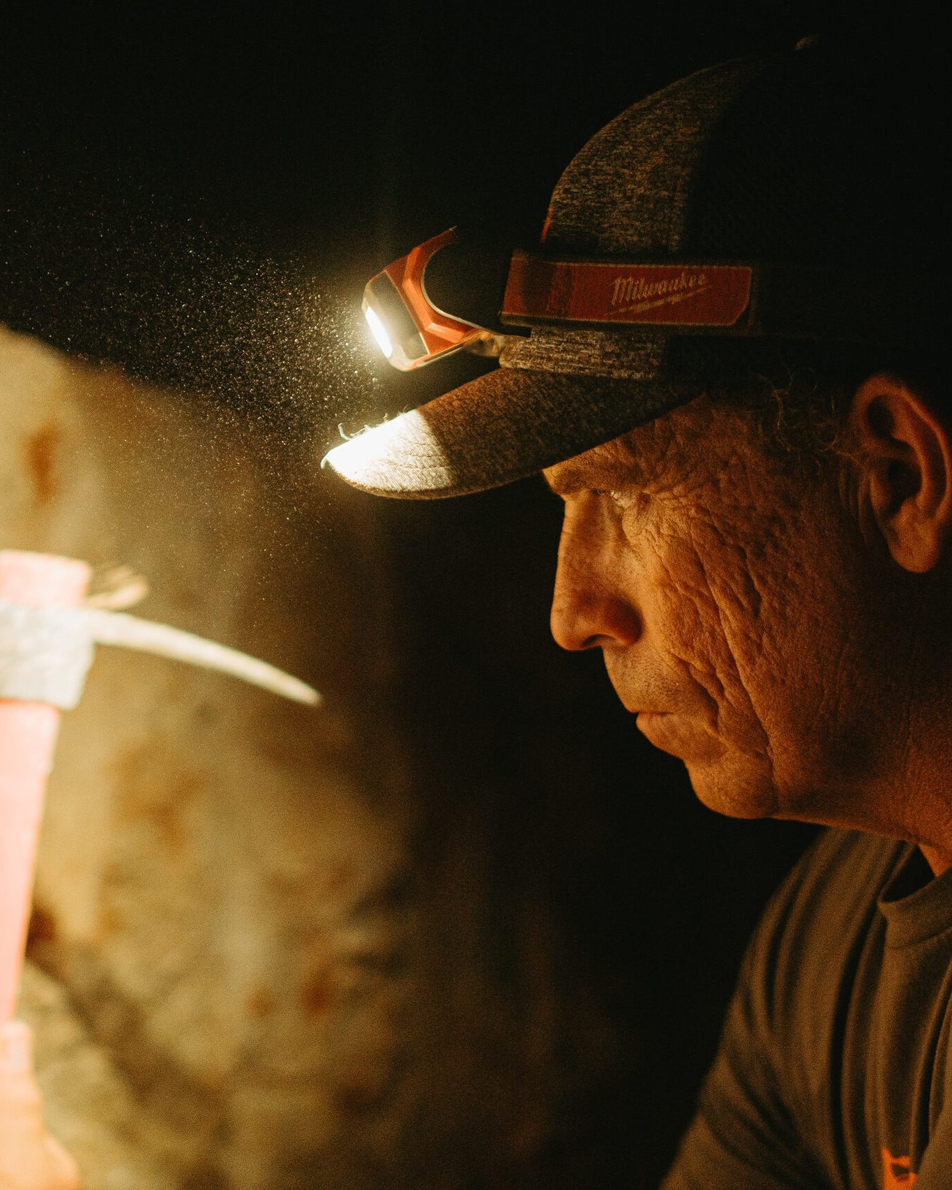 Dan underground in profile, his headlamp lighting the dust as he reads the mine face with his pick raised.