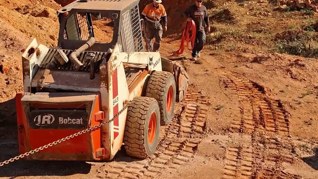 A chained skid steer on the red dirt ramp with two of the crew walking out behind it.