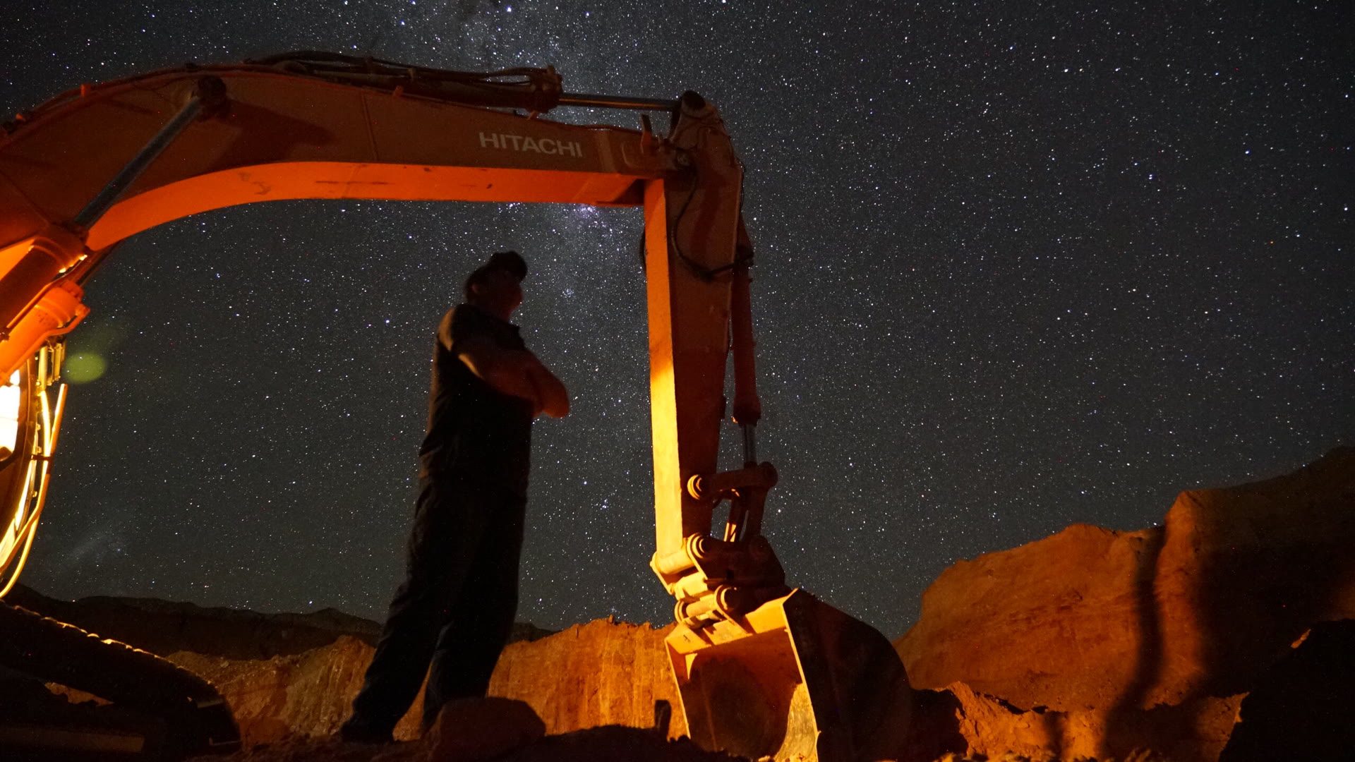 Dan standing under the excavator boom at night, the Milky Way overhead and the work lights on the machine.