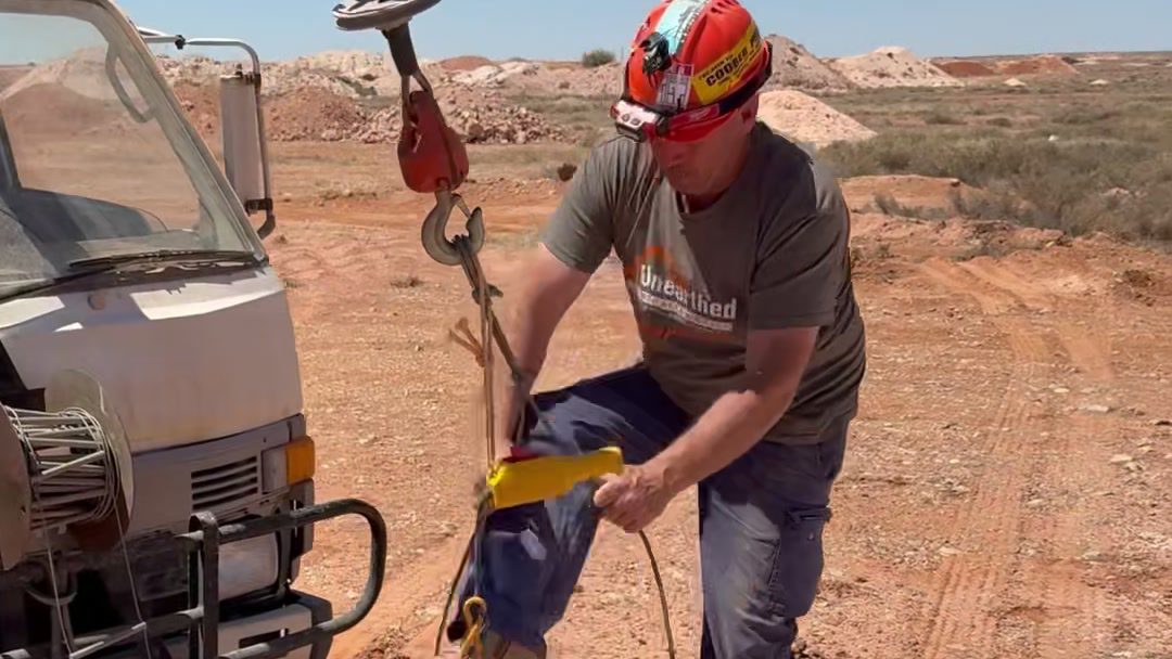 Dan in a hard hat holding the winch hook and cable beside the truck out on the opal field.