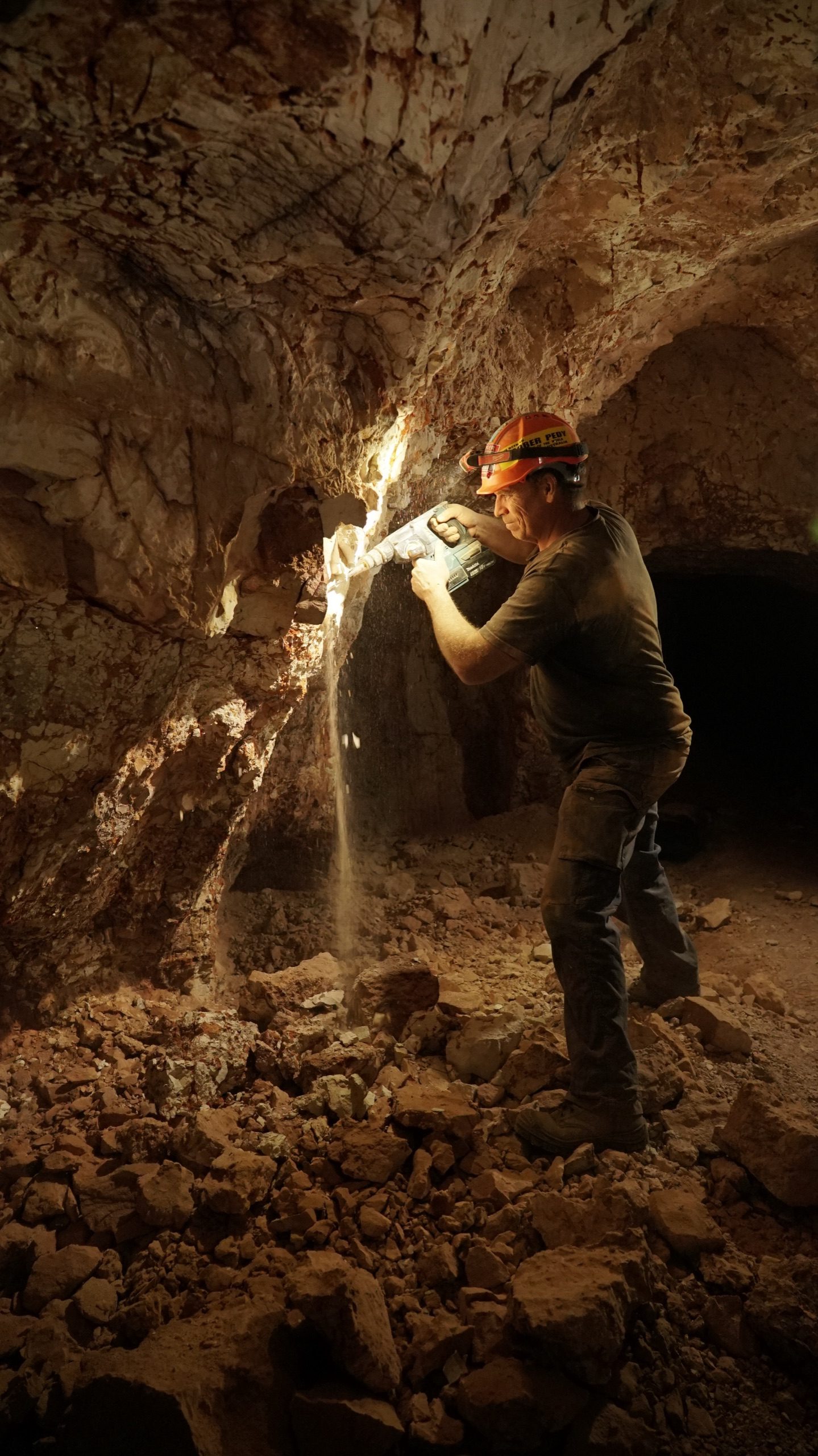 Dan driving the rotary hammer into a lit opal level underground, spoil cascading off the face.