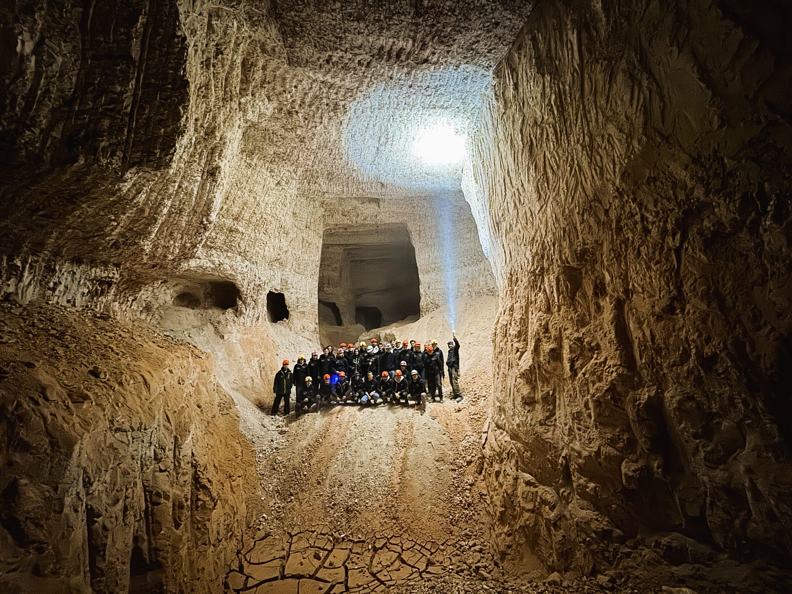 A tour group dwarfed by the walls of a cathedral sized underground chamber, a torch beam pointed up at the roof.